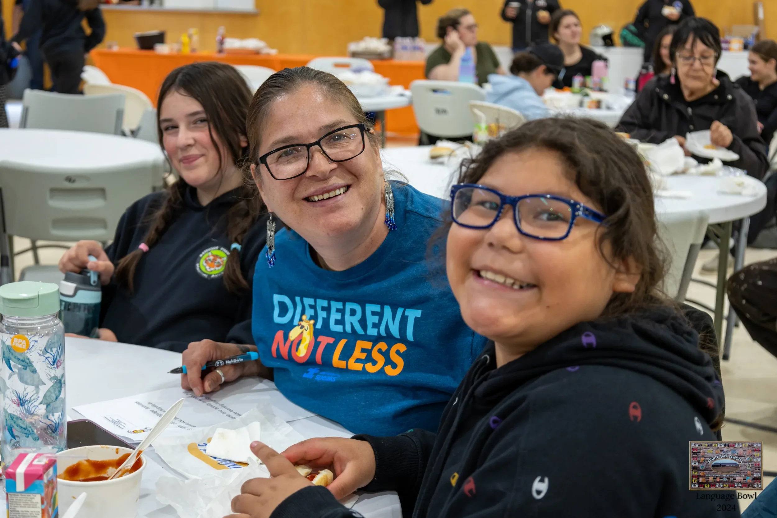 Three smiling people sitting at a table during a group event, with food, drinks, and papers in front of them, in a large indoor space with other people in the background.