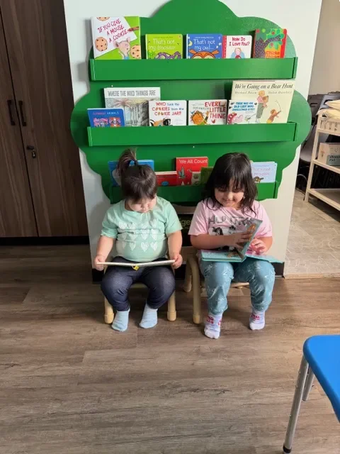 Two young girls sitting in small chairs, reading books in front of a green bookshelf filled with children's books.