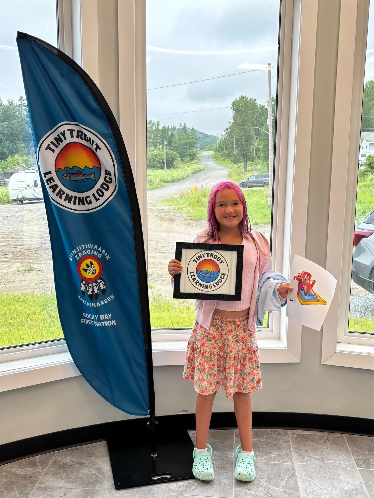 A young girl with purple hair smiling indoors, holding a sign with the Tiny Trout Learning Lodge logo and a paper craft of a boat. Behind her is a window showing a green outdoor landscape with a gravel road and cars. To her left, a blue flag displays the Tiny Trout Learning Lodge logo and other details.
