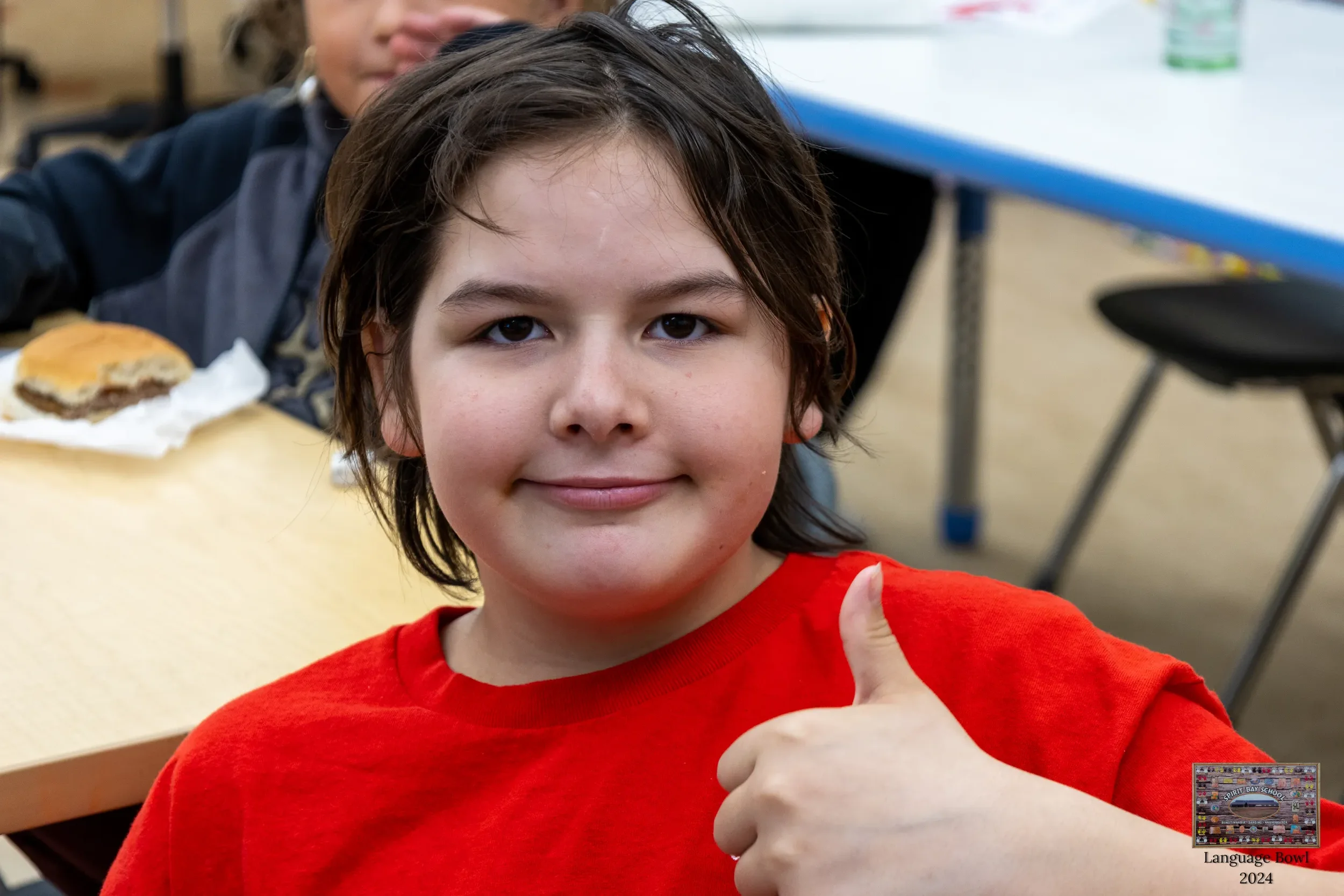 A boy with brown hair and a red shirt giving a thumbs-up inside a classroom.