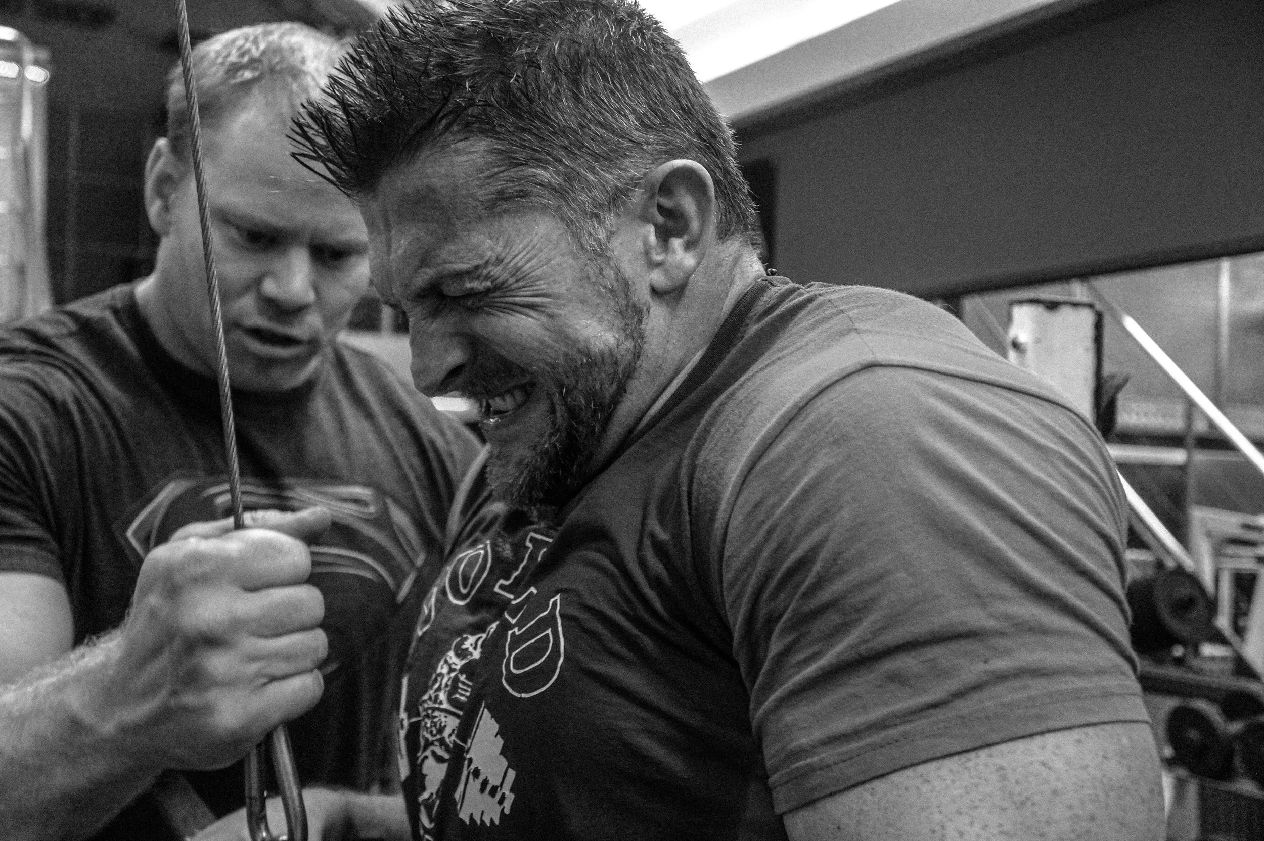Two men at the gym, one with a muscular build and beard, laughing, while working out together, both wearing athletic shirts.