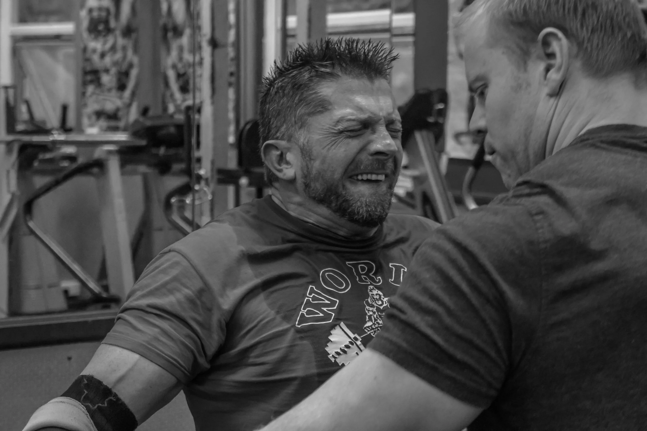 Two men, one older with a beard smiling and eyes closed, and one younger with short hair, facing each other in a gym, mid-exercise or interaction.
