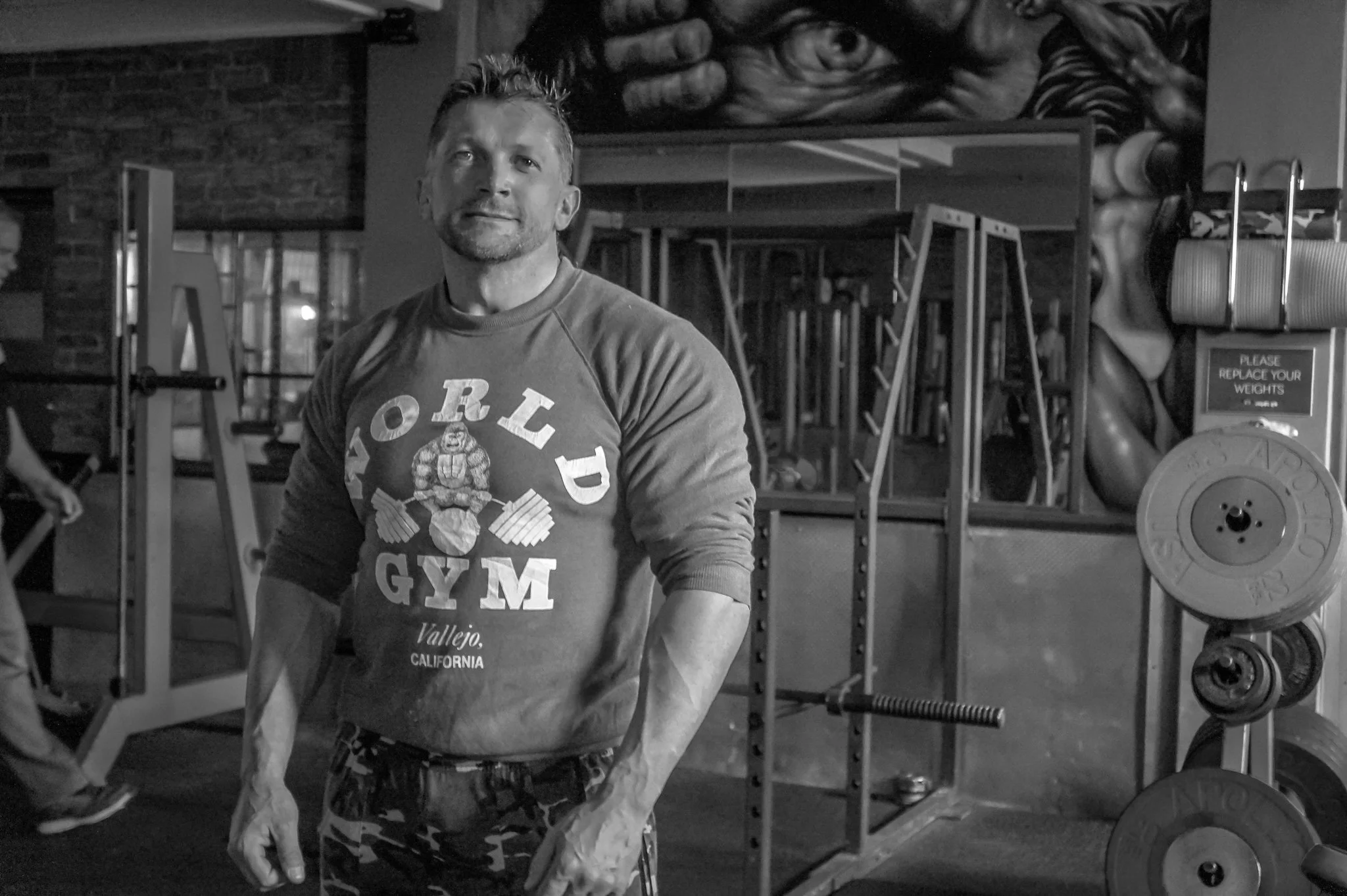 A muscular man standing in a gym, wearing a hoodie that says "World Gym Vallejo, California," with various gym equipment and a mural of a face on the wall behind him.