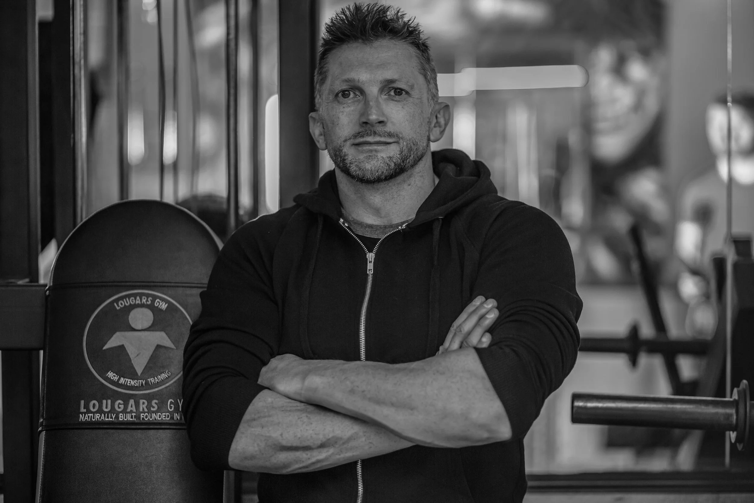 A man with crossed arms and a serious expression standing inside a gym, with gym equipment in the background.