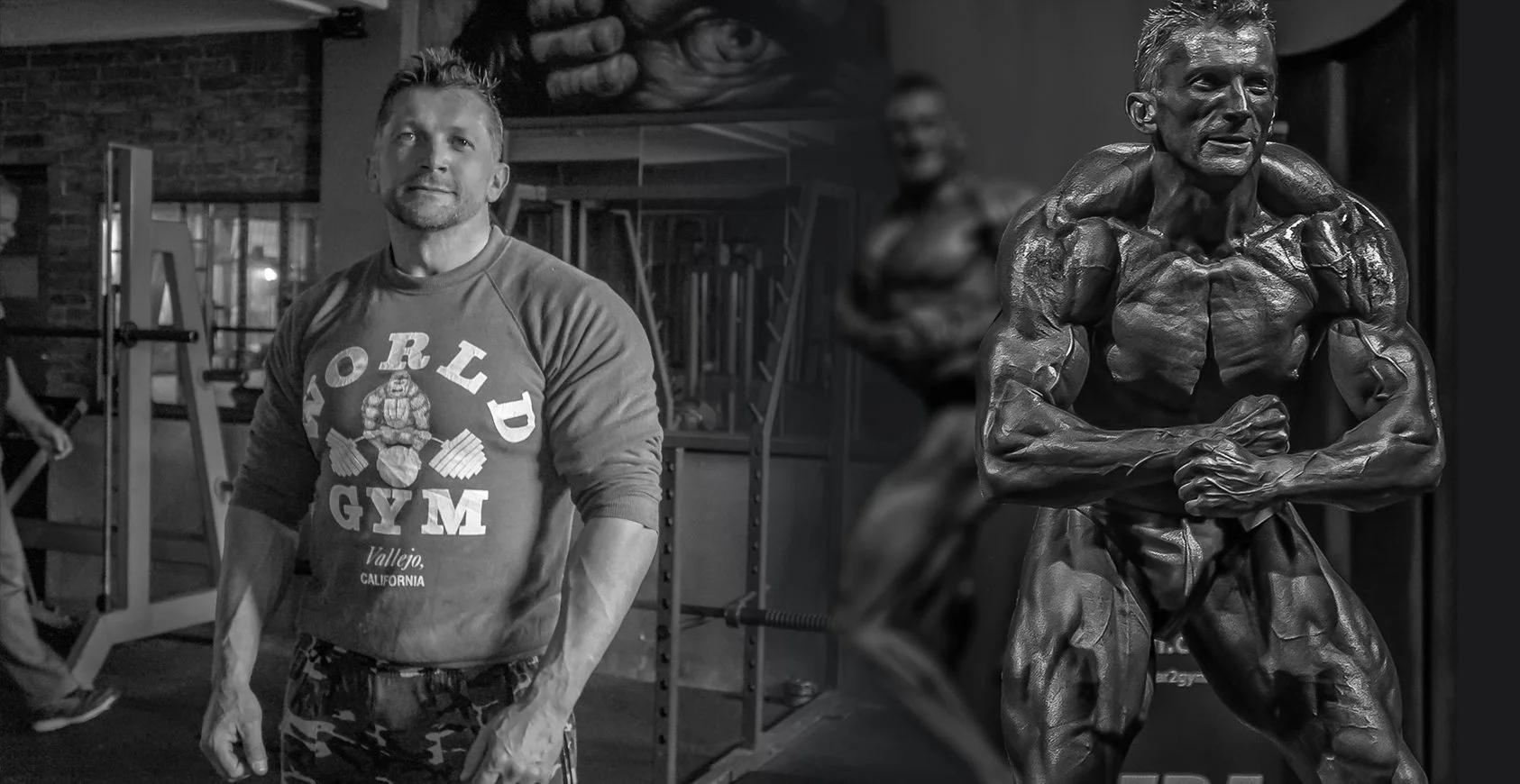 A man in a Gym shirt standing in a gym with exercise equipment and a mural in the background, next to a statue of a muscular man flexing.