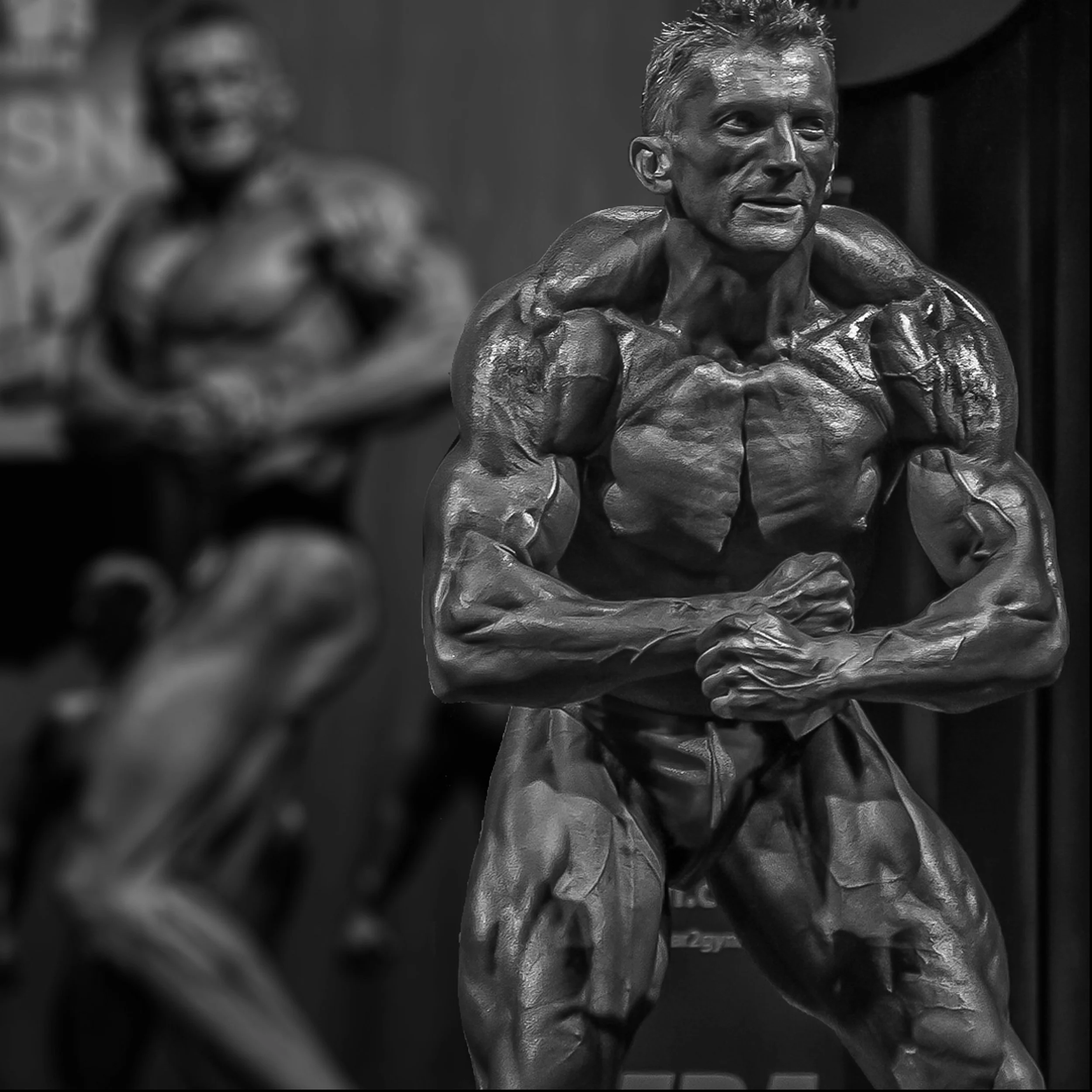Black and white photo of a wax statue of a male bodybuilder flexing muscles in a bodybuilding pose, with another blurred bodybuilder statue in the background.