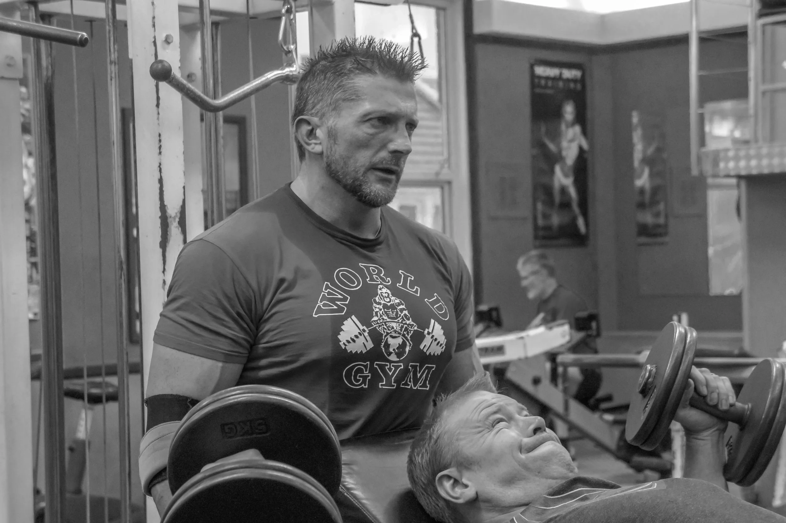 A man in a 'World Gym' T-shirt assists an elderly man lifting dumbbells while lying on a workout bench at a gym.