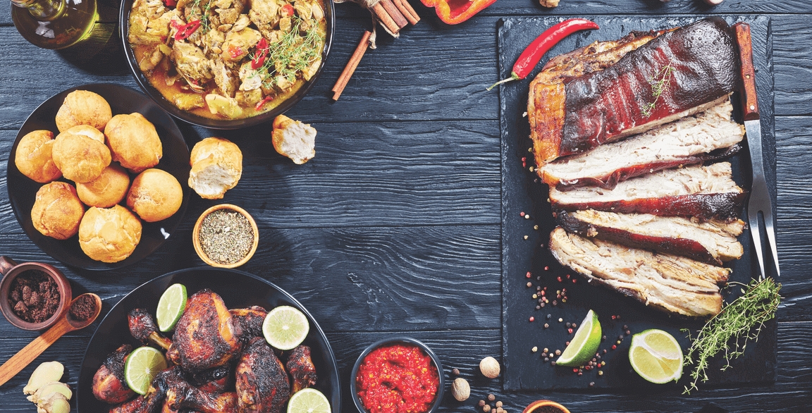 A spread of food including sliced smoked pork ribs with lime, a bowl of curry, fried balls, grilled chicken wings with lime, and various sauces and spices on a dark wooden table.