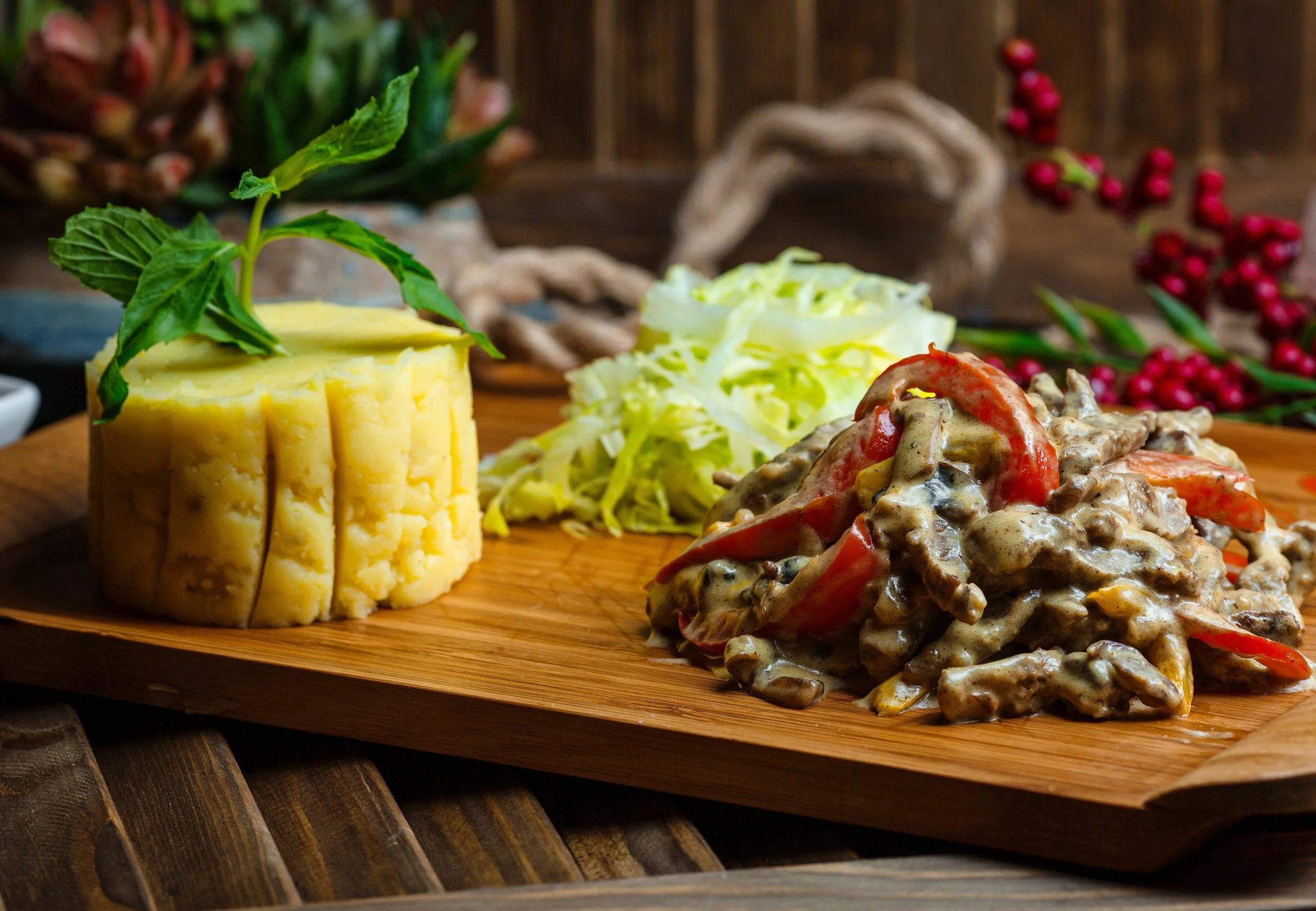 A wooden platter with mashed potatoes garnished with mint, shredded lettuce, and a beef and vegetable stew with red bell peppers.