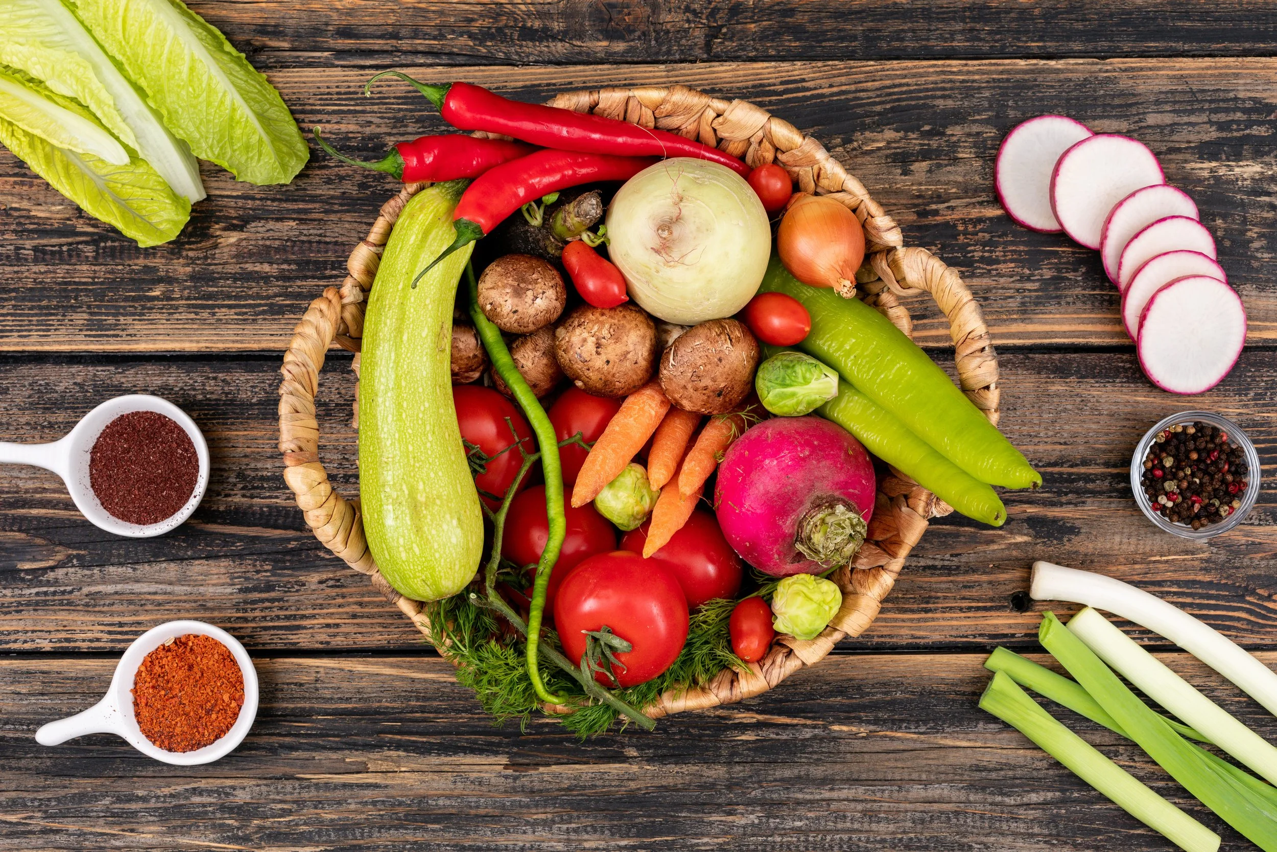 A basket of fresh vegetables including tomatoes, carrots, radishes, cucumbers, potatoes, green chili, onion, radicchio, and a round white squash, with additional vegetables and spices arranged on a rustic wooden surface.