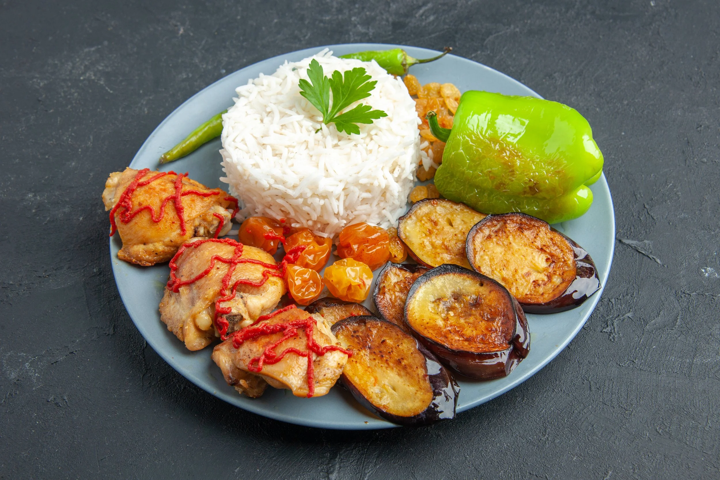 Plate of white rice garnished with parsley, grilled eggplant slices, roasted green peppers, and cooked chicken pieces with ketchup drizzle, accompanied by yellow cherry tomatoes, green chili pepper, and rice on a blue plate on dark background.