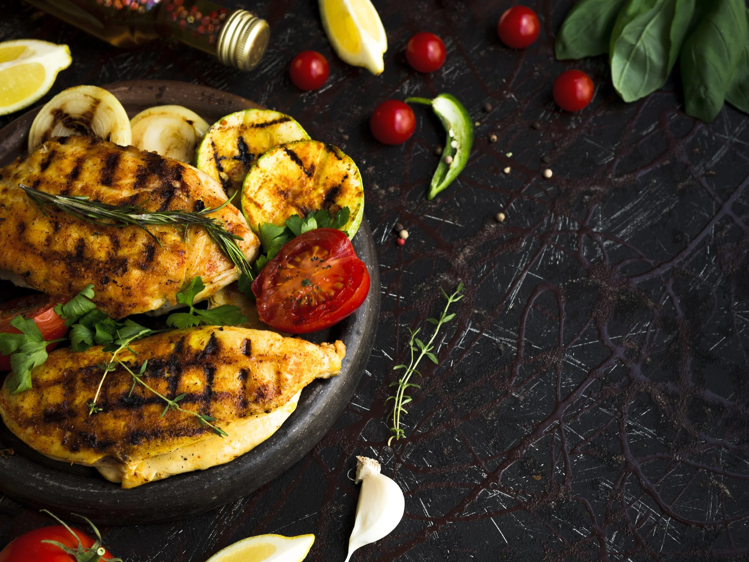 Grilled chicken breast with herbs, cherry tomatoes, zucchini, onion, lemon wedges, basil, garlic, and pepper on a dark textured surface.