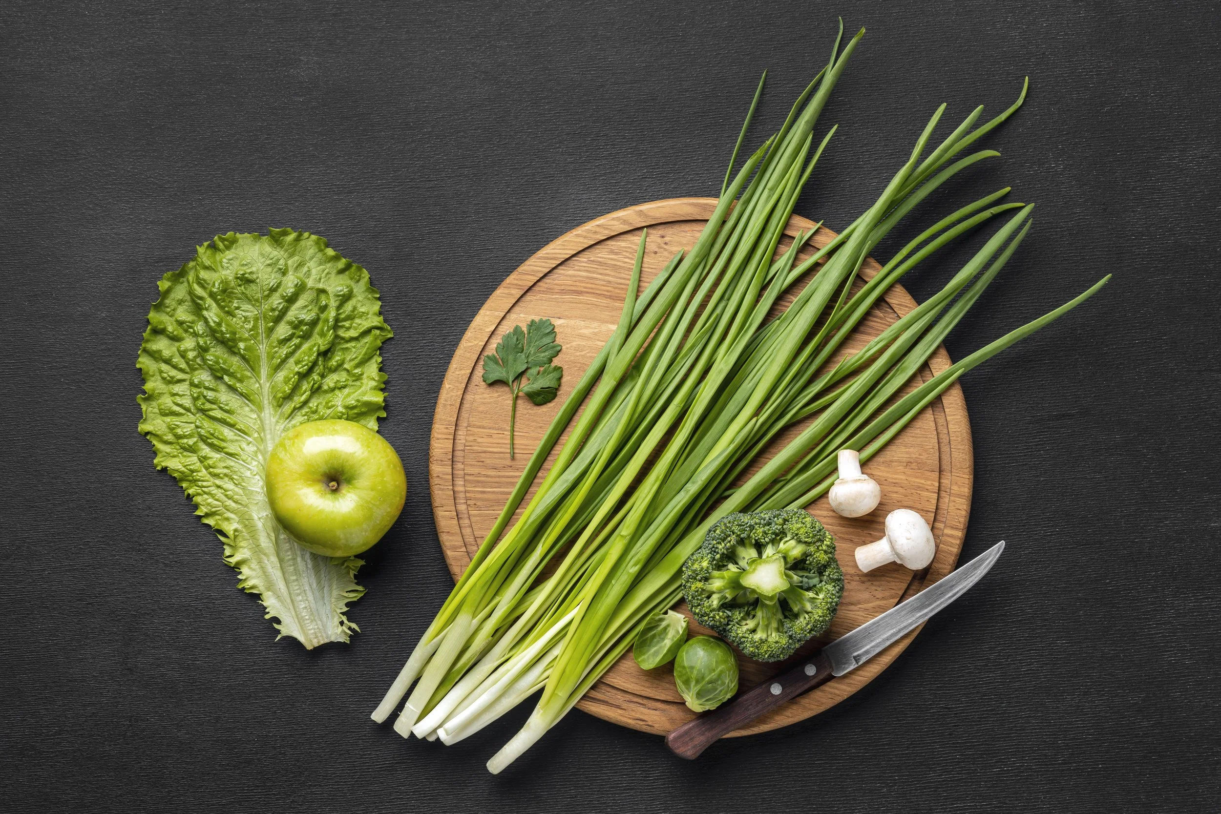 Fresh green vegetables and herbs on a round wooden cutting board, including green onions, broccoli, mushrooms, Brussels sprouts, and a leaf of lettuce, with a green apple and cilantro leaf on a black surface.