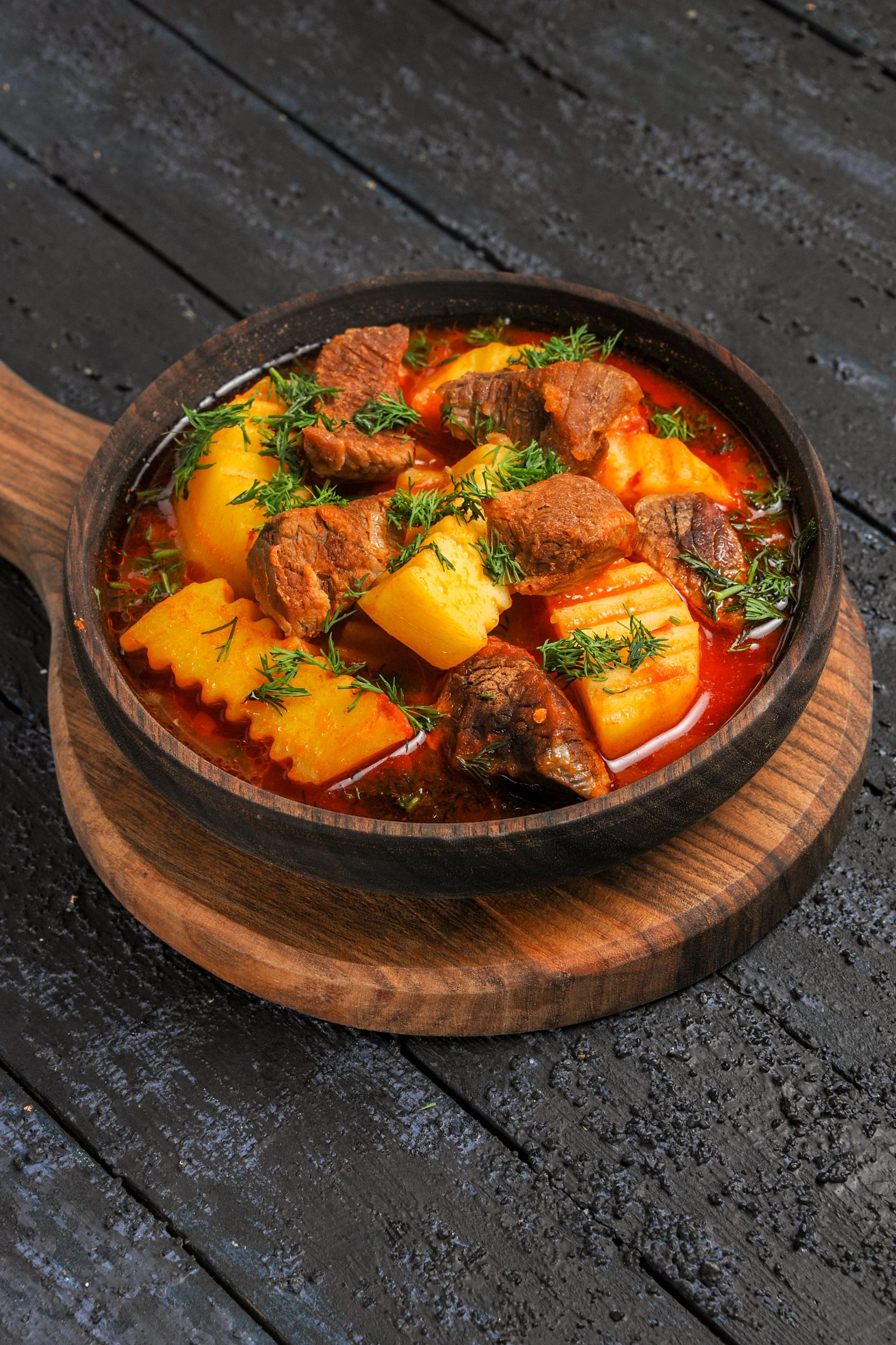 A bowl of beef stew with potatoes, carrots, and herbs on a wooden tray.