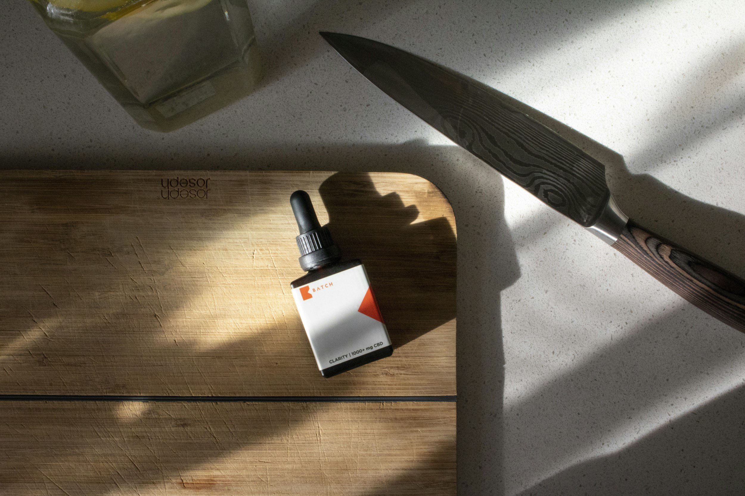 A kitchen countertop with a wooden cutting board, a small bottle of CBD oil, a chef's knife, and part of a glass jar or container with lemon slices.