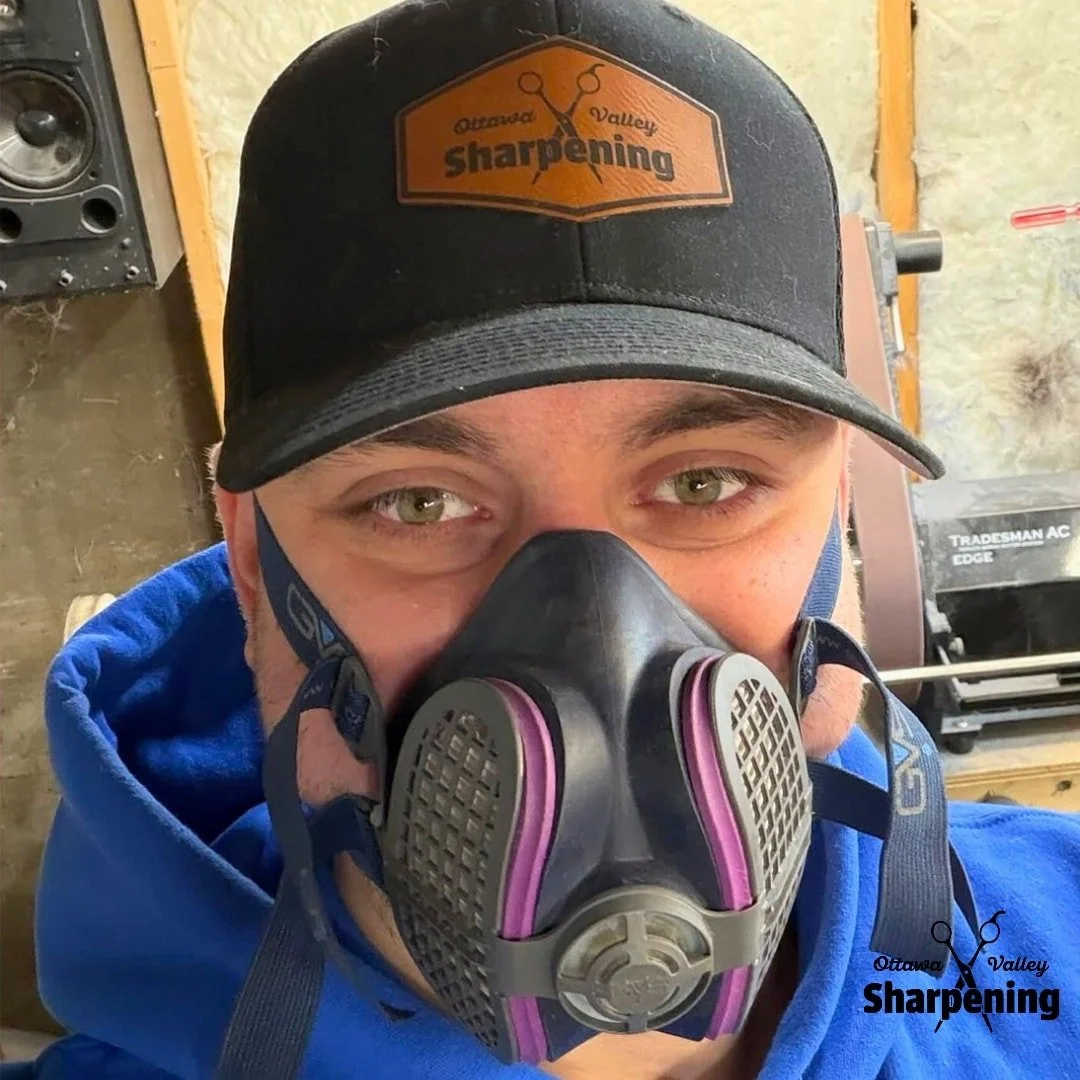 A man wearing a black cap with an orange logo that reads 'Ottawa Valley Sharpening' and a black face mask with purple accents. The background reveals a workshop setting with tools and equipment.