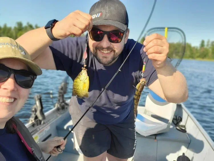 Two people on a boat holding fishing rods with small fish caught on the hooks, smiling and showing their catch in a sunny outdoors setting.