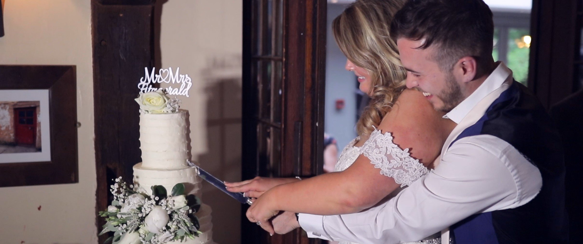 A bride and groom cutting a wedding cake together, smiling, with a wedding topper that reads 'Mr & Mrs Fitzgerald'.