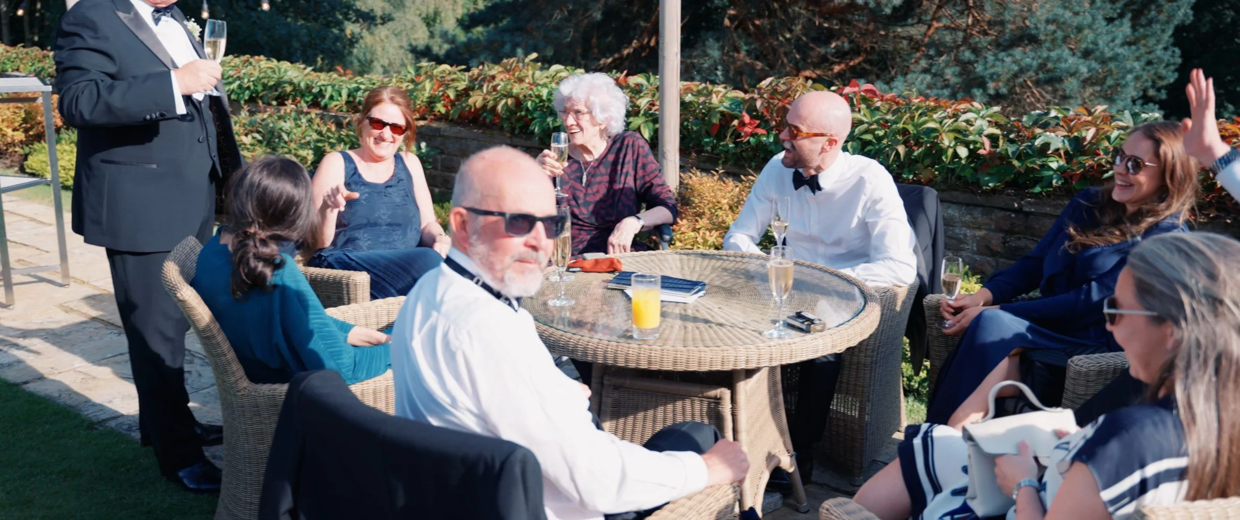 People enjoying a garden party outdoors, sitting at a round wicker table with drinks, with a person in a black suit standing nearby.