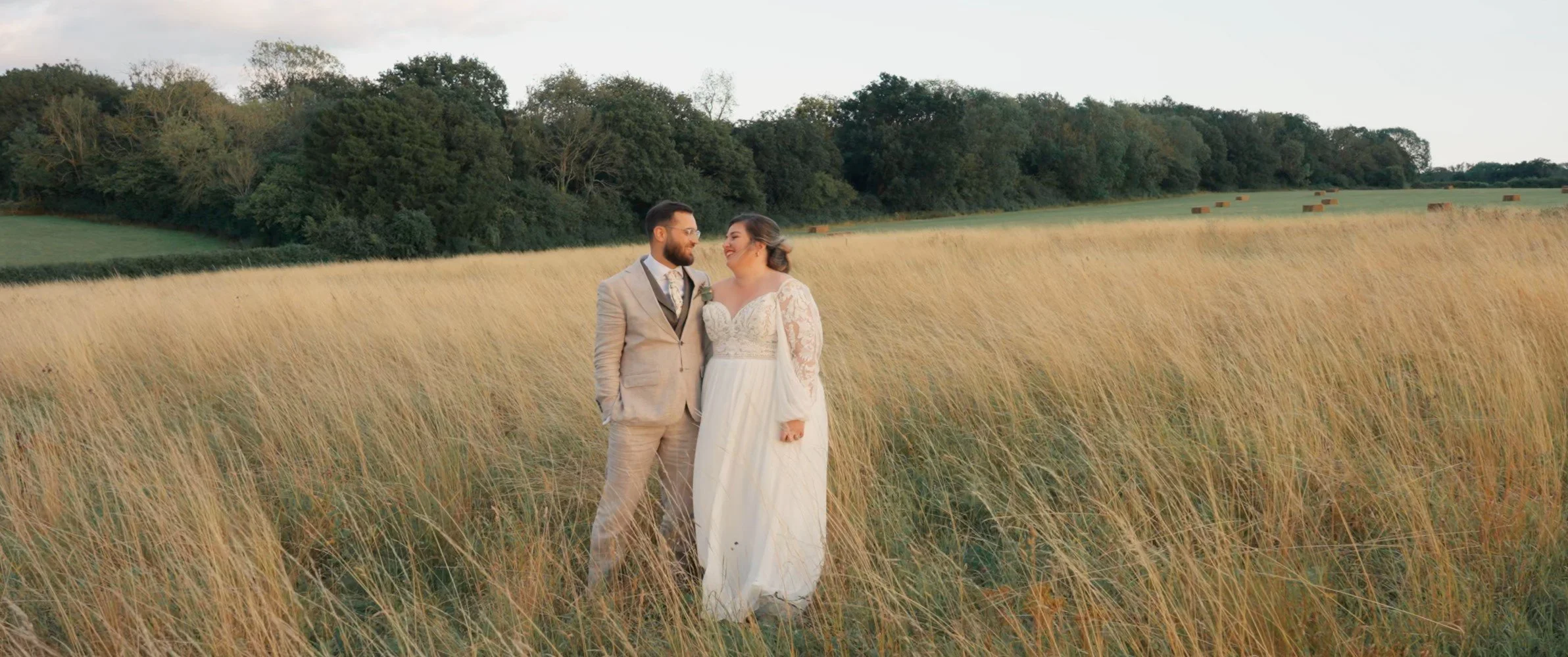 A bride and groom in wedding attire standing in a field of tall, golden grass, smiling and looking at each other with trees and hay bales in the background during sunset.