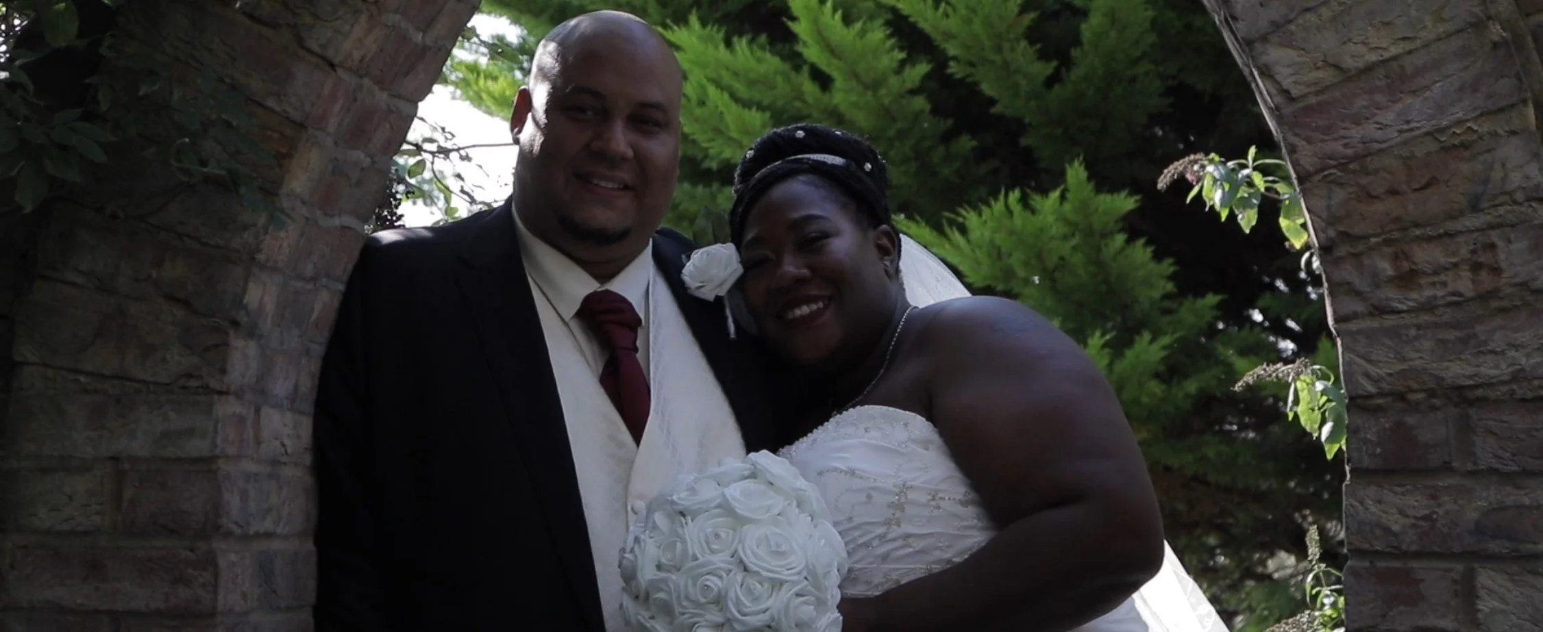 A newlywed couple stands close together outdoors, framed by a stone archway with green trees in the background. The groom is wearing a suit with a maroon tie, and the bride is in a white wedding dress holding a bouquet of white roses, smiling.