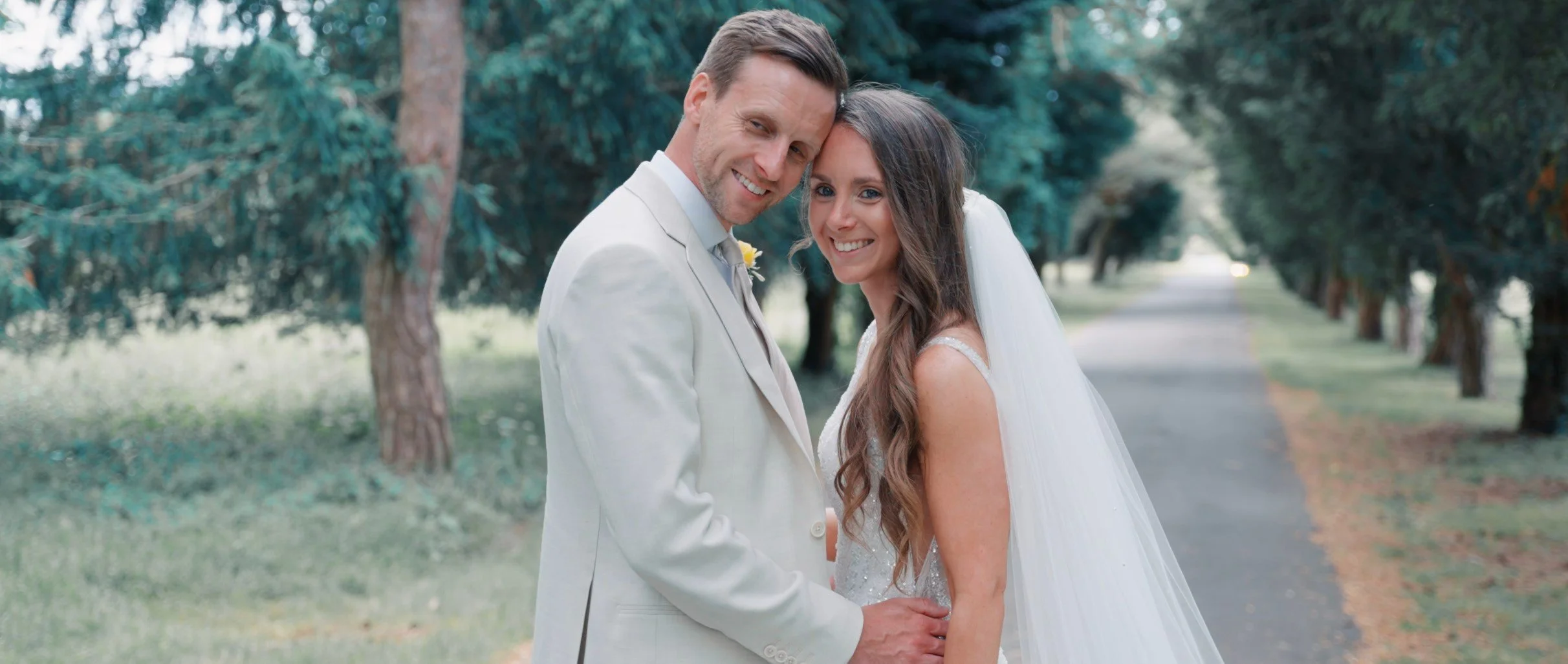 A smiling bride and groom standing close together outdoors on a tree-lined path, celebrating their wedding day.