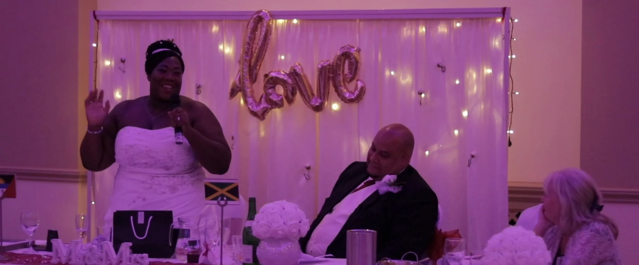 Woman in white dress speaking into a microphone at a celebration with a decorated backdrop and people seated at a table.