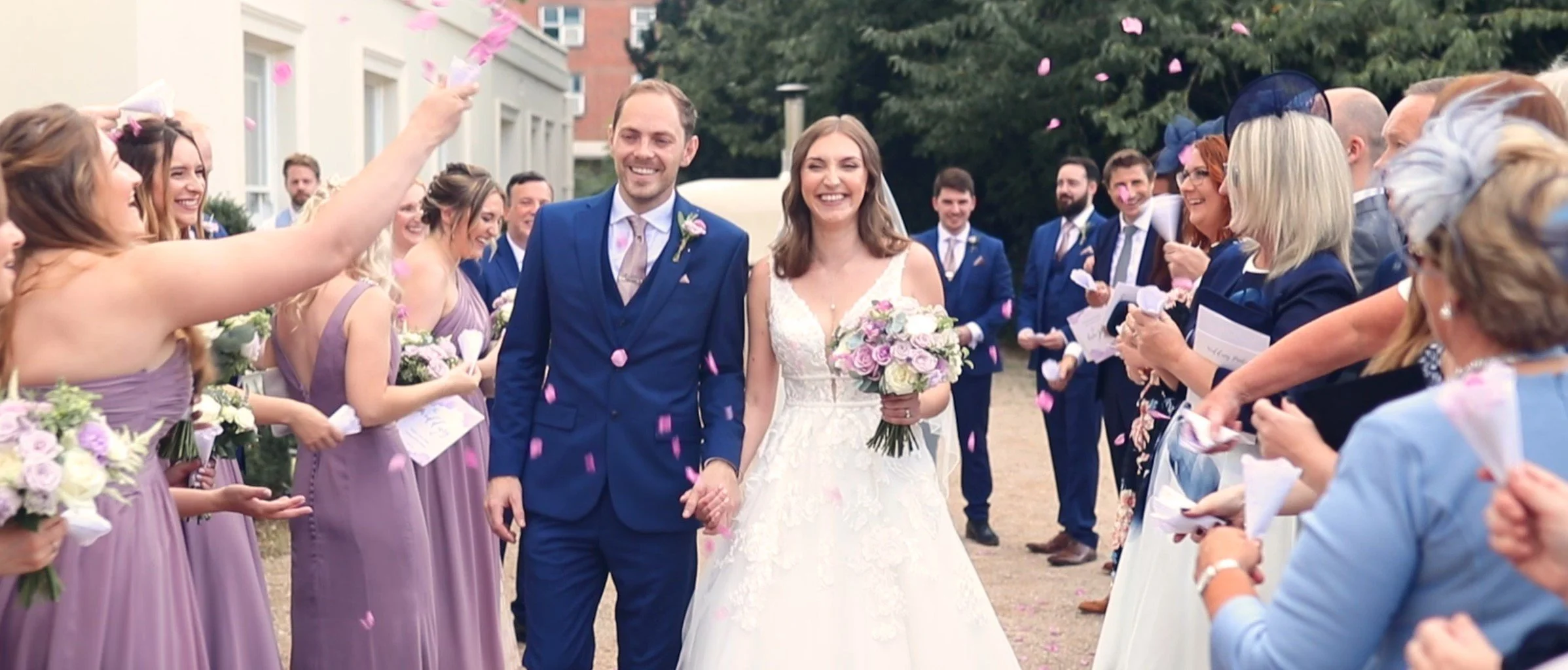 A bride and groom smiling as they walk hand-in-hand through a crowd of wedding guests outdoors, with pink flower petals falling around them.