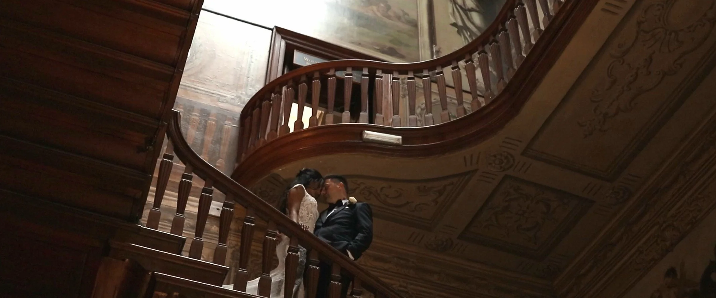 A couple dressed in formal wedding attire sharing a kiss on a grand wooden staircase with ornate ceiling details in the background.