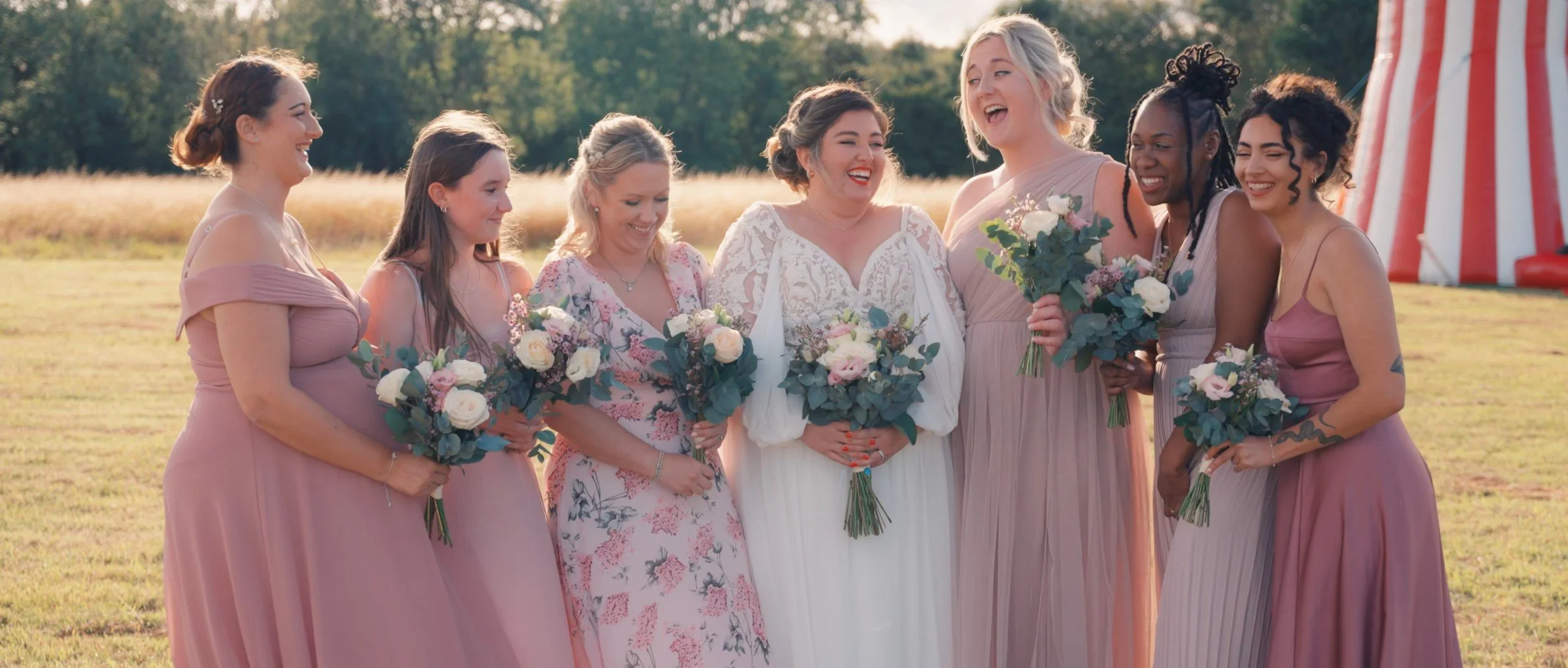 A group of women, dressed in pastel and floral dresses, smiling and holding bouquets of flowers, stands outdoors in a field with trees in the background, celebrating a wedding.