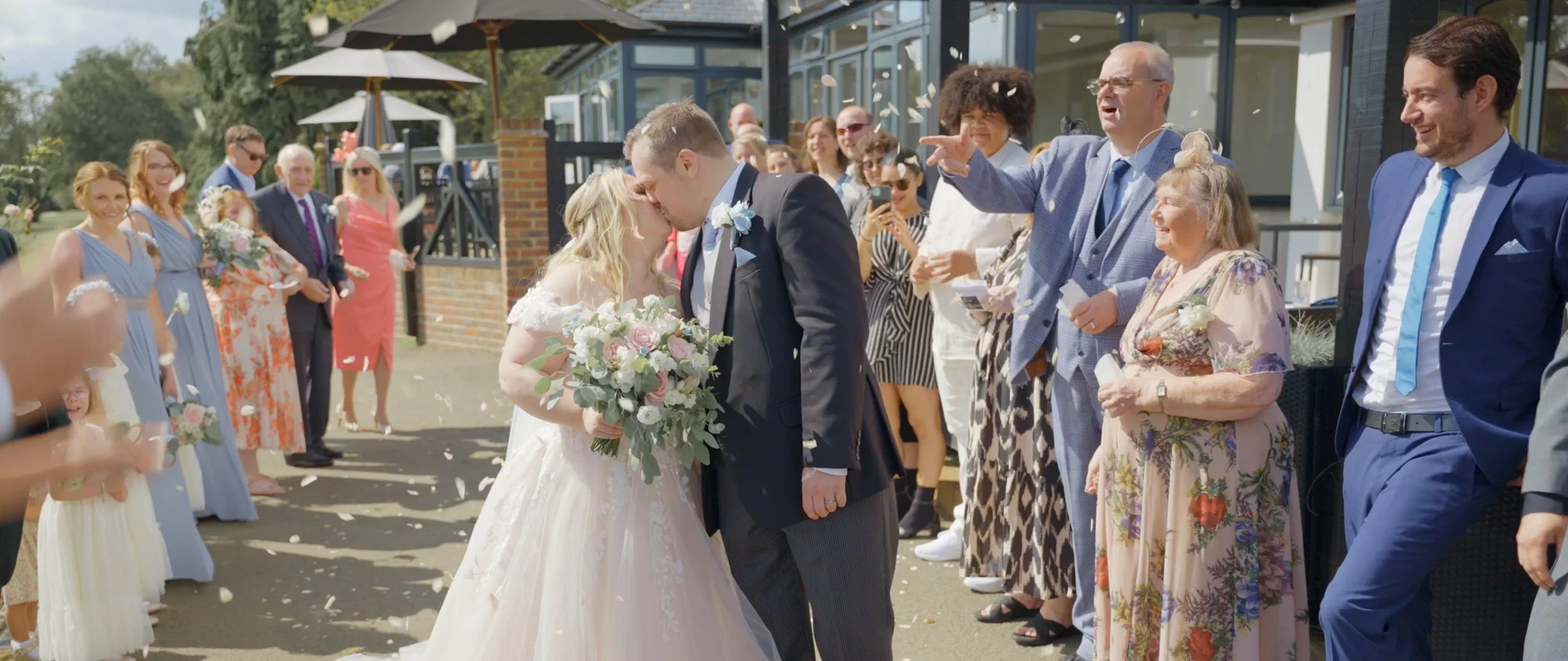 A newlywed couple kisses during their outdoor wedding celebration, surrounded by family and friends who are throwing flower petals and smiling.