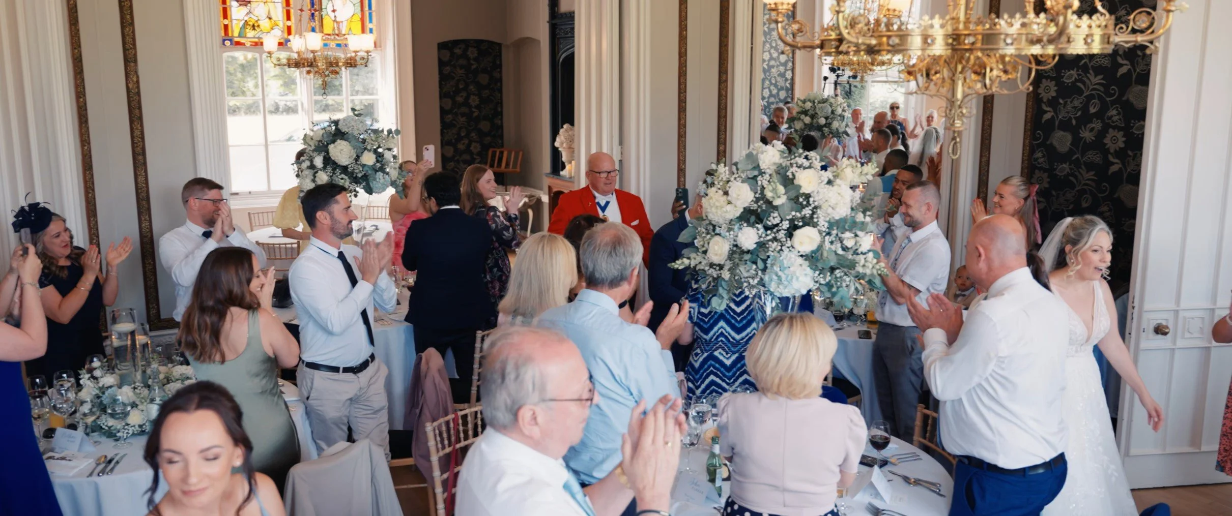 Guests at a wedding reception standing and clapping in a decorated banquet hall with floral centerpieces and chandeliers.