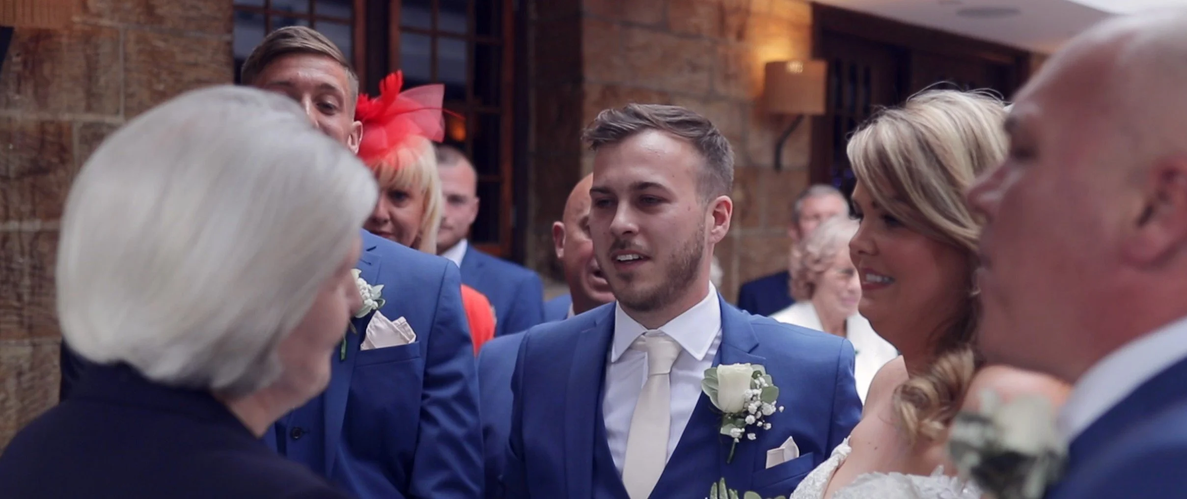 People at a wedding ceremony, including a woman with white hair, a man in a blue suit, a woman with blonde hair, and a man with a shaved head, standing indoors in a rustic setting.