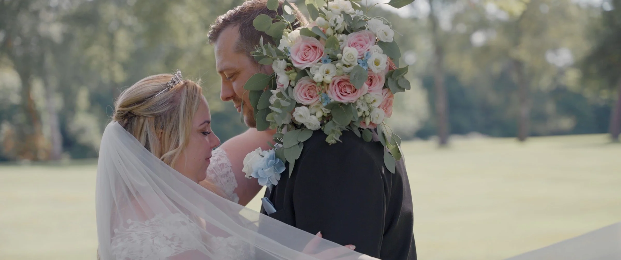 A bride and groom embrace during a wedding ceremony outdoors in a park-like setting, with the bride wearing a veil and the groom carrying a bouquet of pink and white flowers with green leaves.