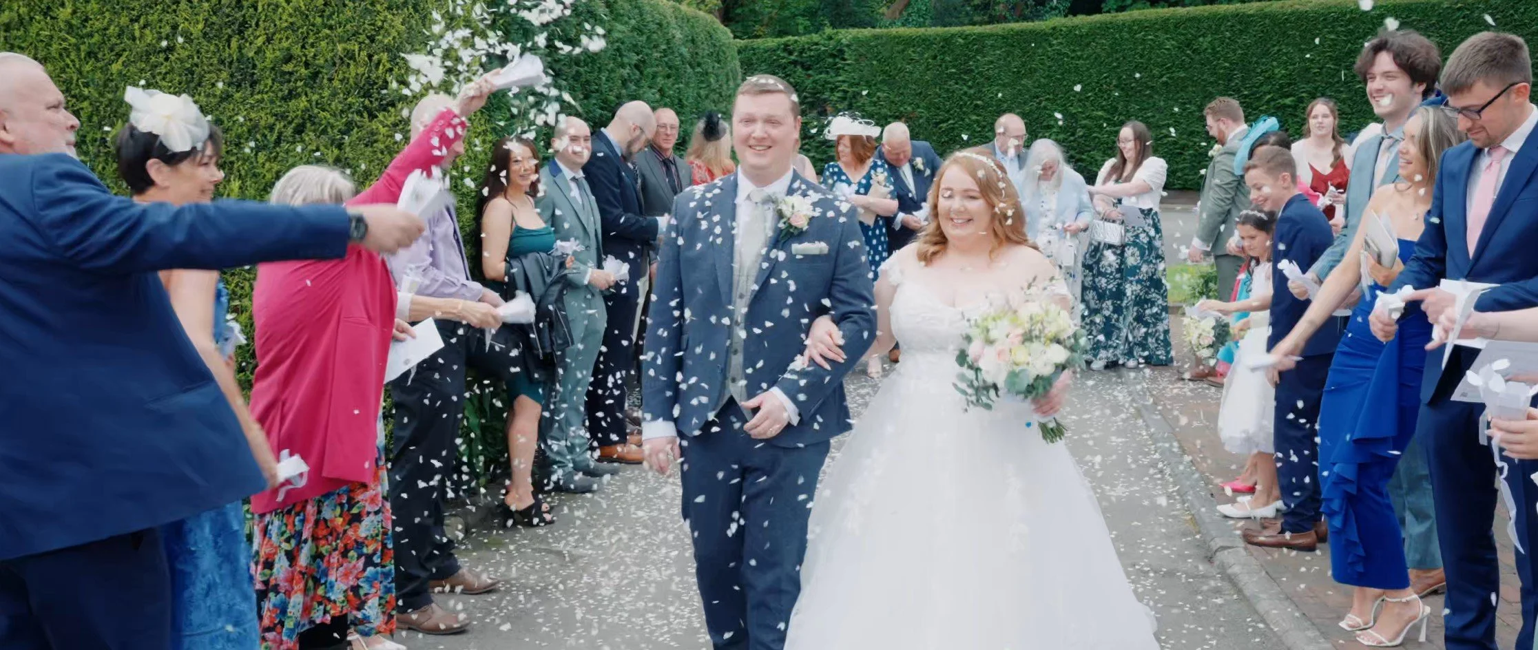 A newlywed couple in wedding attire walking through a pathway lined with guests, surrounded by greenery, as guests throw confetti in celebration.