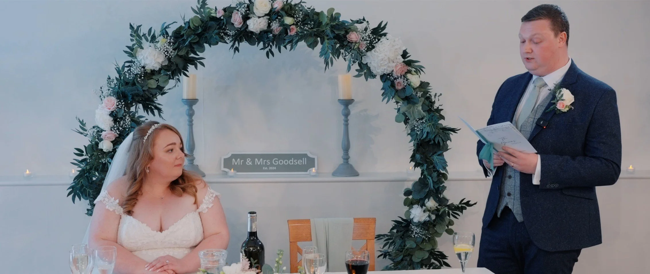 Bride and groom listening to vows during their wedding ceremony in front of a floral arch with candles and a Mr & Mrs sign in the background.