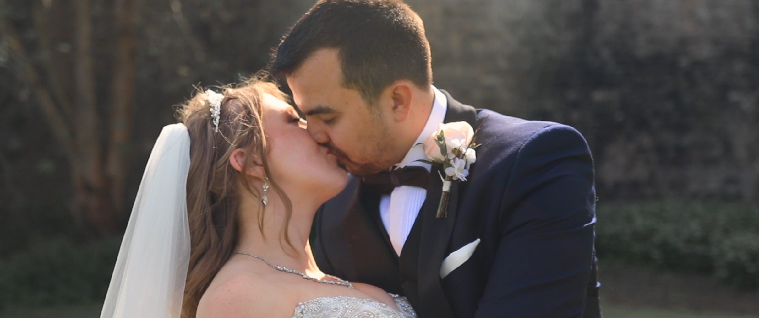 A bride and groom sharing a kiss outdoors during their wedding, with the bride wearing a veil and wedding dress and the groom in a suit with a boutonniere.