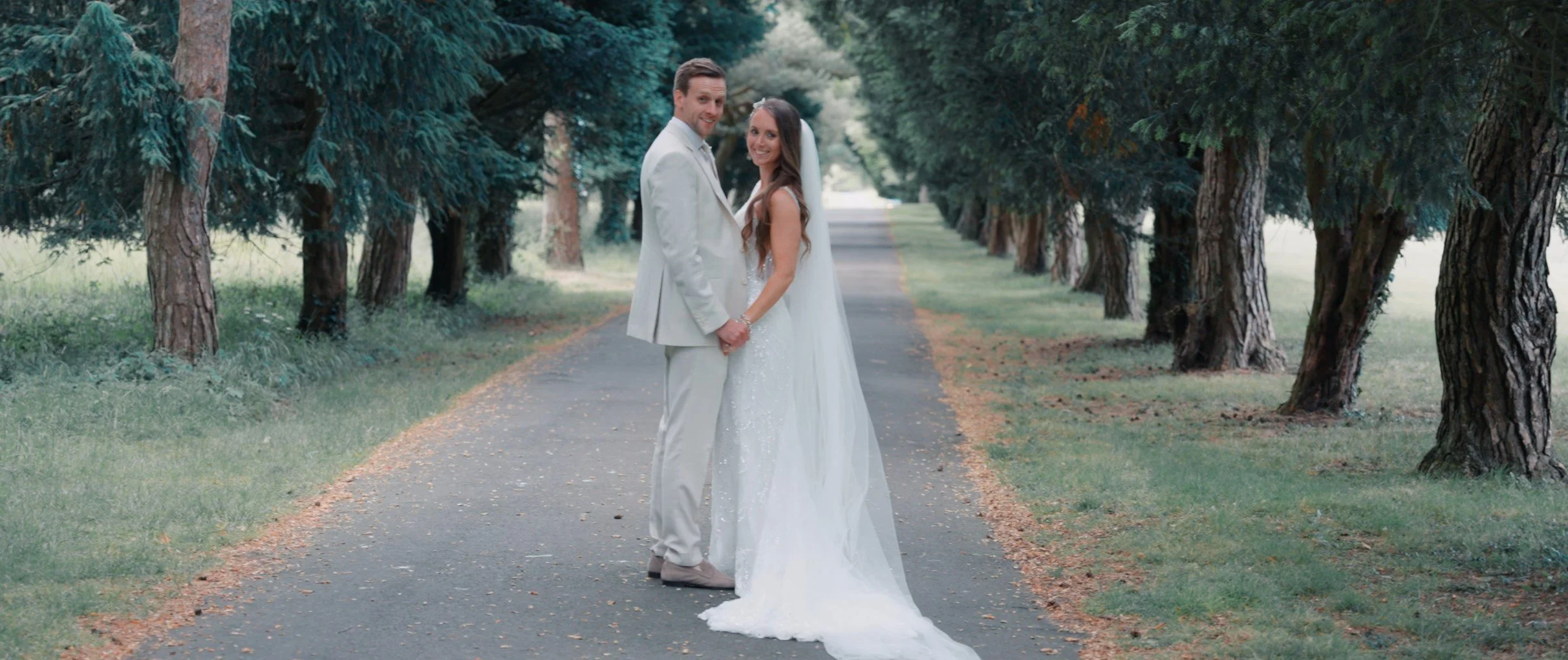 A newlywed couple standing hand-in-hand on a tree-lined path, smiling in wedding attire.