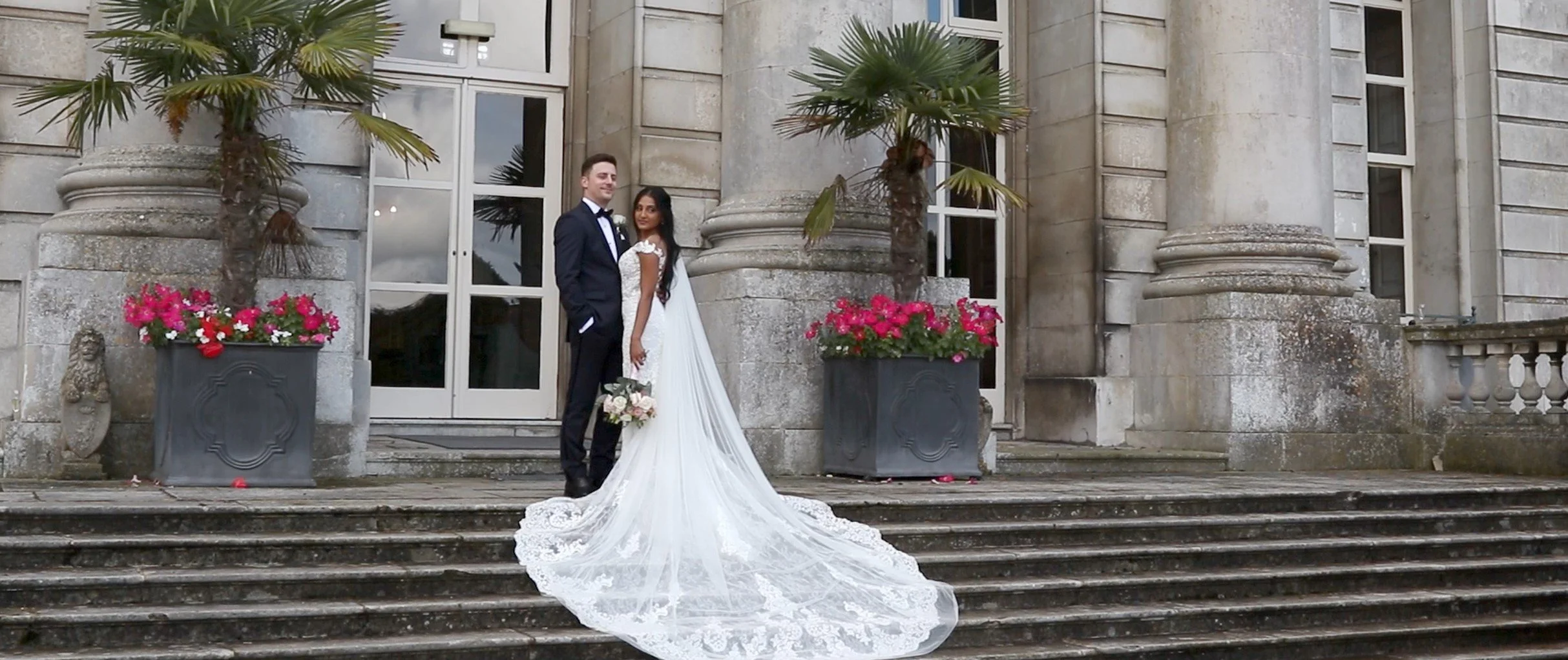 A bride and groom standing on front steps of a grand stone building with large glass doors, floral planters, and palm trees, celebrating their wedding.