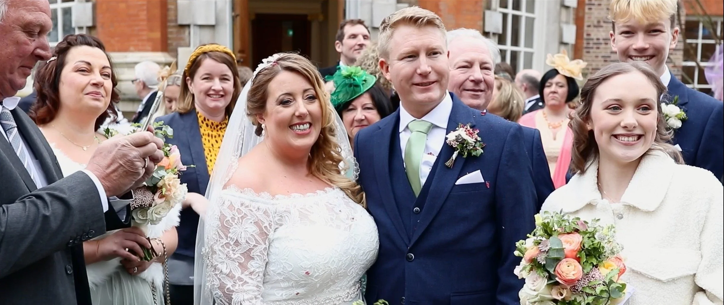 Group of people at a wedding, with the bride and groom smiling in the center, surrounded by family and friends, with theatrical hats and floral bouquets.