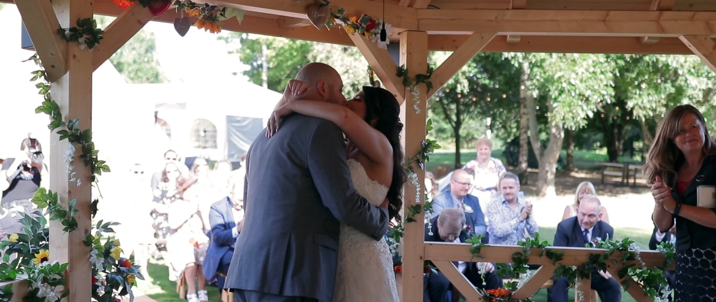 A bride and groom are kissing under a wooden archway decorated with greenery and flowers, during their outdoor wedding ceremony. Guests are seated and standing in the background under trees, watching and celebrating.