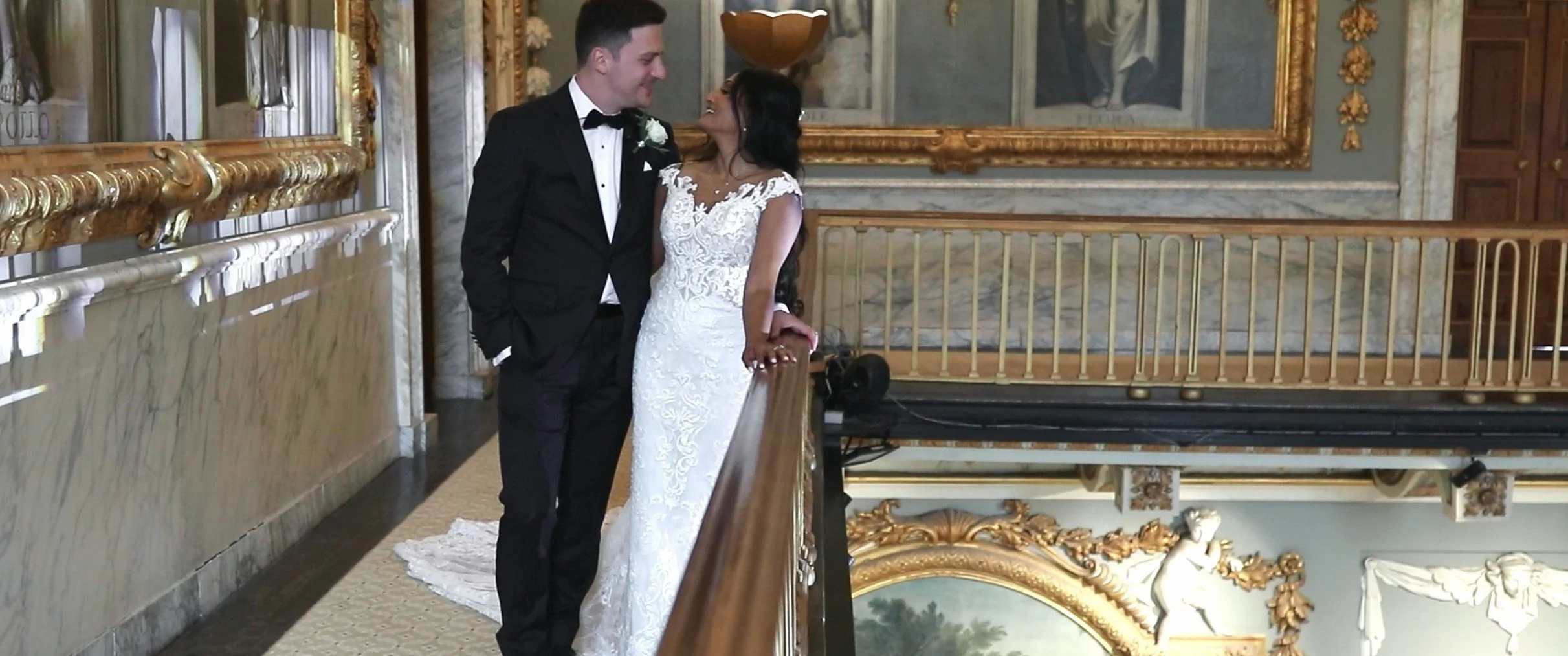 A newlywed couple stands close together inside an opulent, decorated hall. The groom wears a black tuxedo with a bow tie, and the bride wears a white lace wedding gown. They are smiling and gazing at each other, with the bride resting her hand on a r
