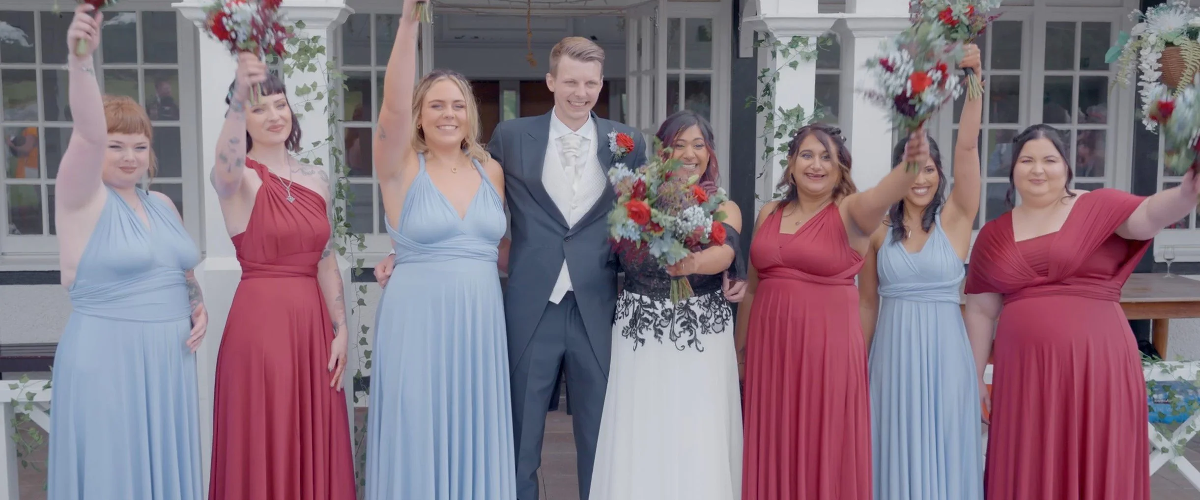 Wedding party standing together outside a venue, with some women holding bouquets and raising them outwards, celebrating with the bride and groom in the center.