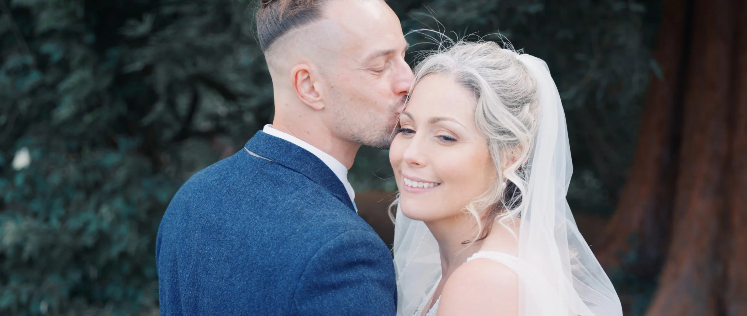 A groom kissing a bride on the forehead during a wedding ceremony outdoors, with green foliage in the background.
