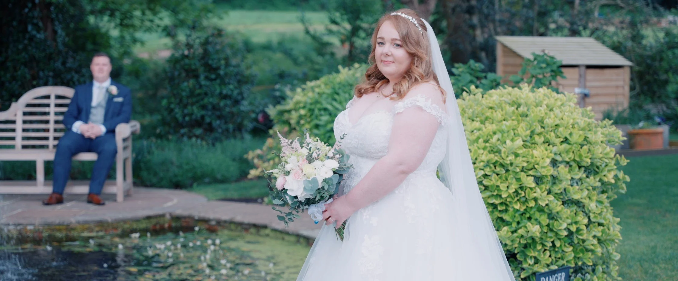 A bride in a white wedding dress holding a bouquet of flowers standing outdoors with a green garden, a pond, and a seated man in a suit in the background.