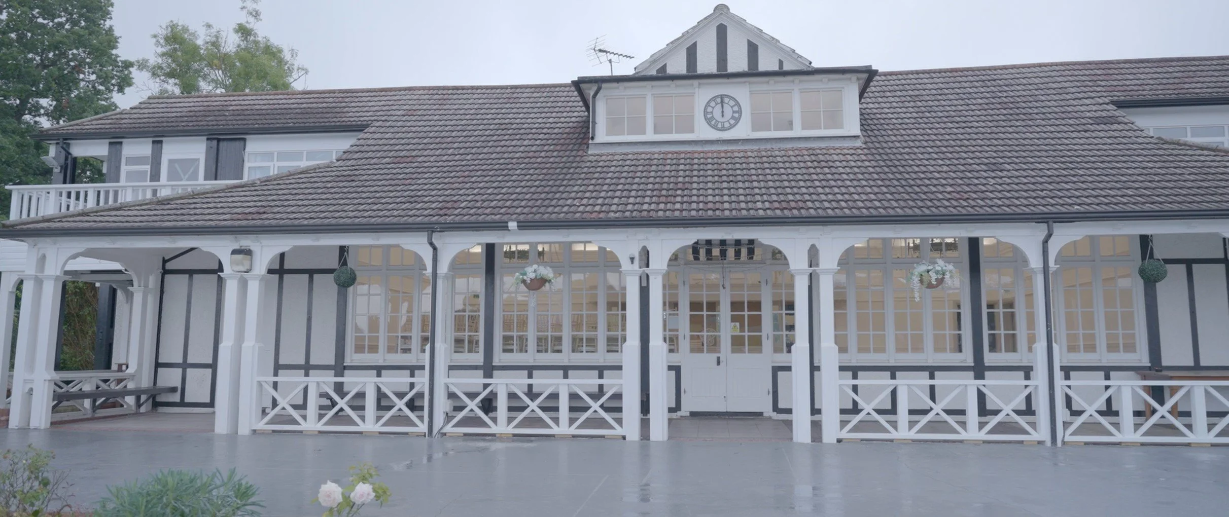 White building with black trim and a clock on the upper section, surrounded by an enclosed porch with white railing and hanging flower baskets, and a tiled roof.