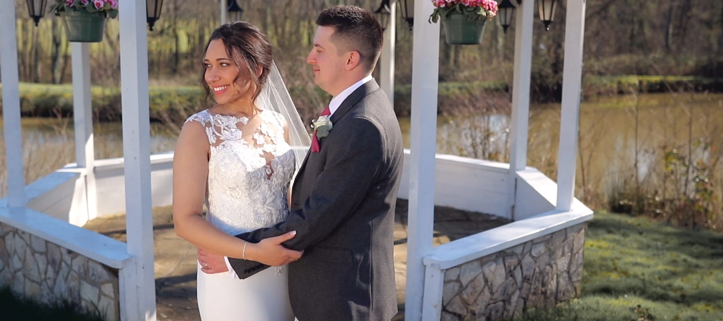 A bride and groom standing close together outdoors in front of a white gazebo, holding hands and smiling at each other. The bride is wearing a white lace wedding gown and veil, and the groom is wearing a dark gray suit with a red tie. There are hangi