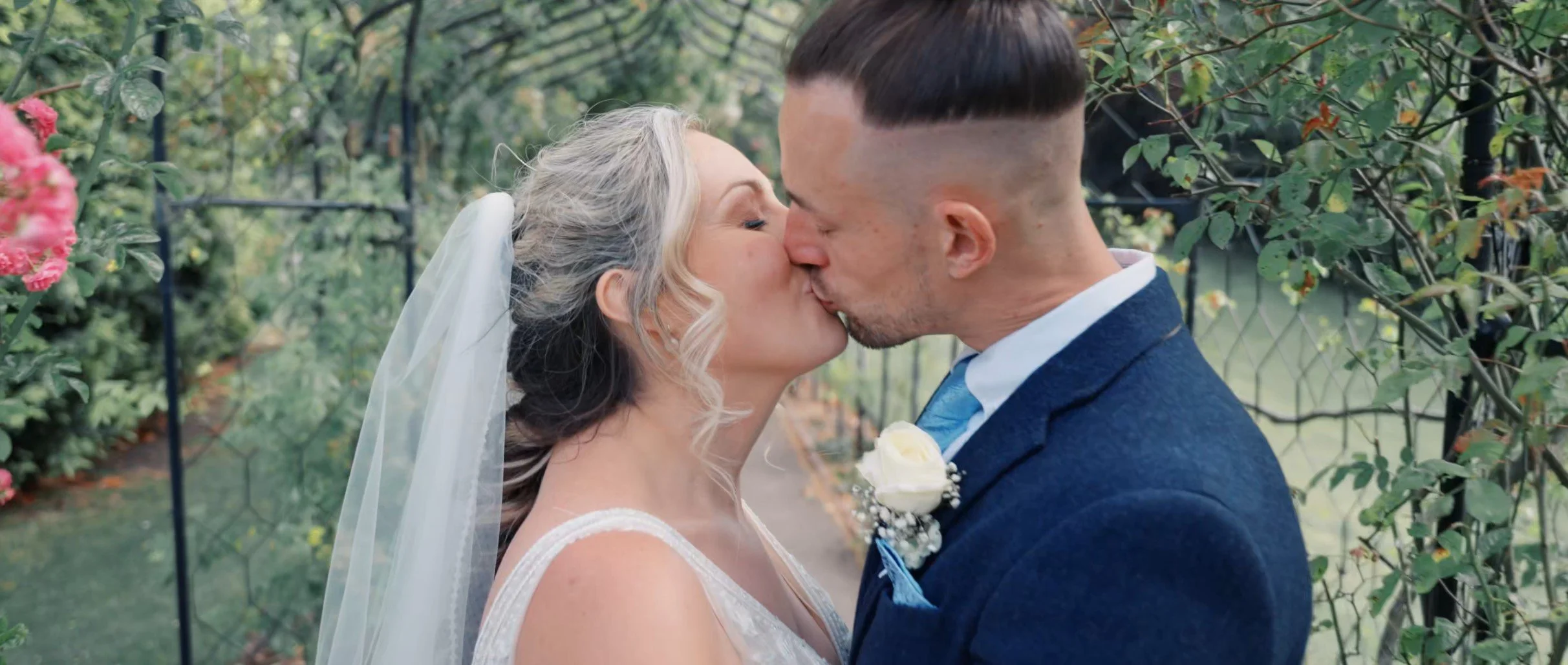 A bride and groom kissing outdoors in a garden setting, with flowers and greenery in the background.