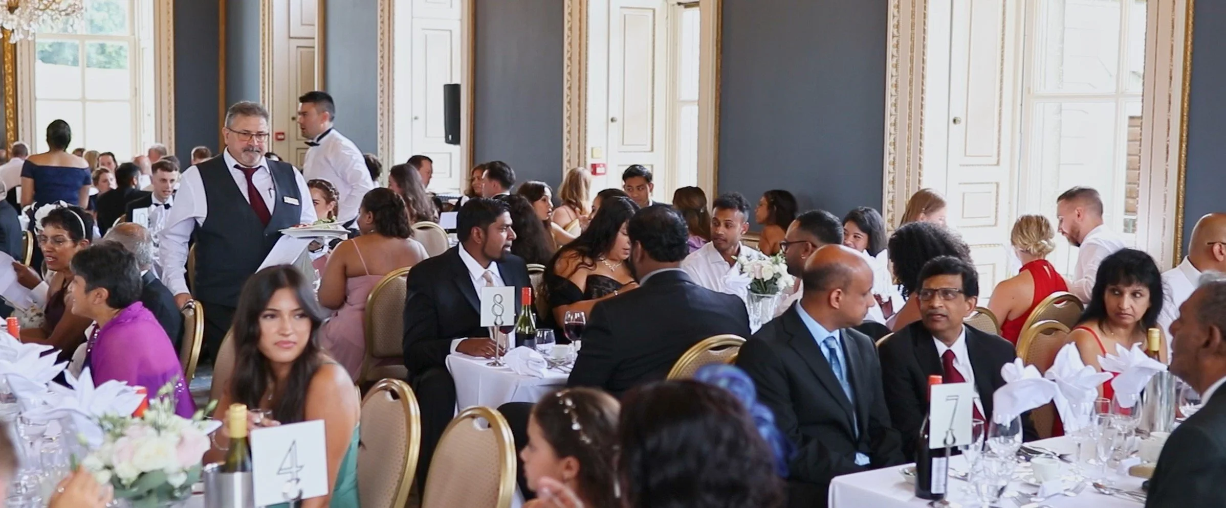Guests dining at round tables in an elegant banquet hall with large windows and ornate gold trim.