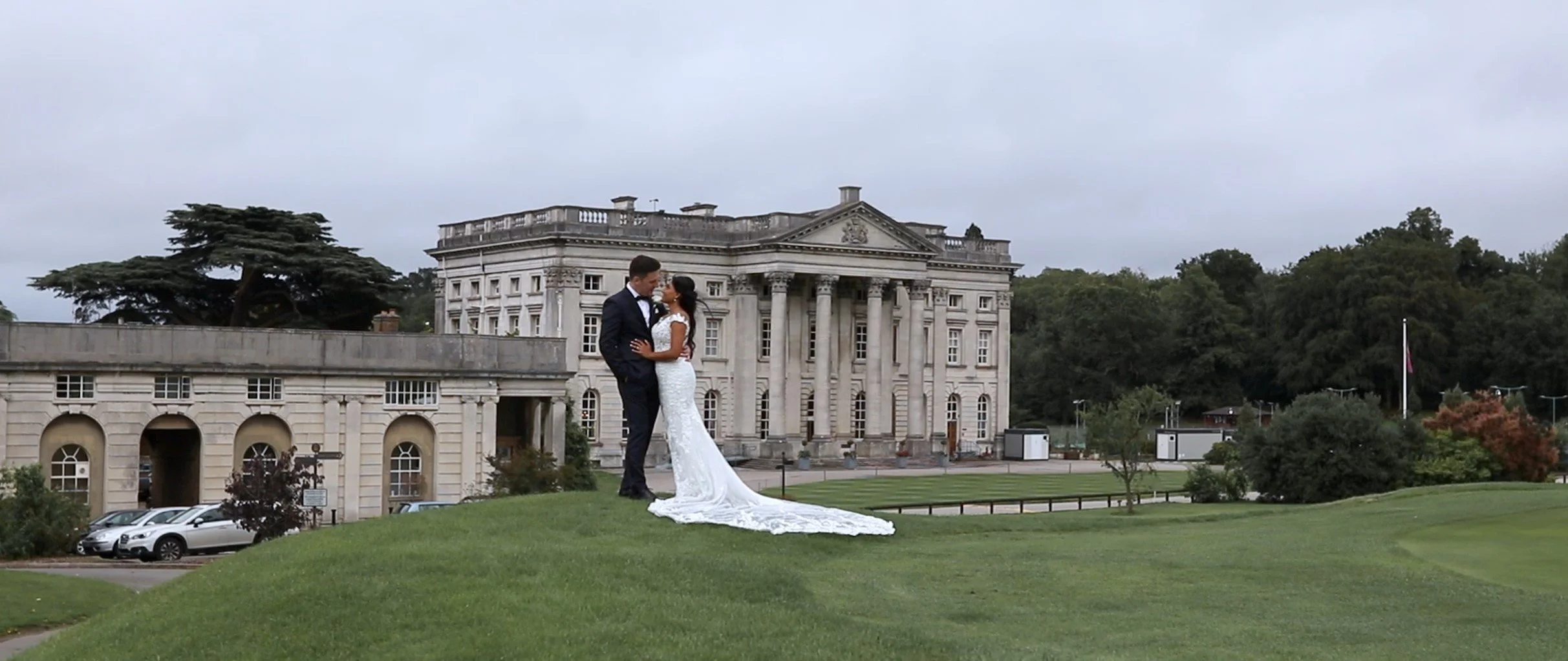 A bride and groom standing on a grassy hill, kissing with a large historic building behind them, surrounded by trees and parked cars.